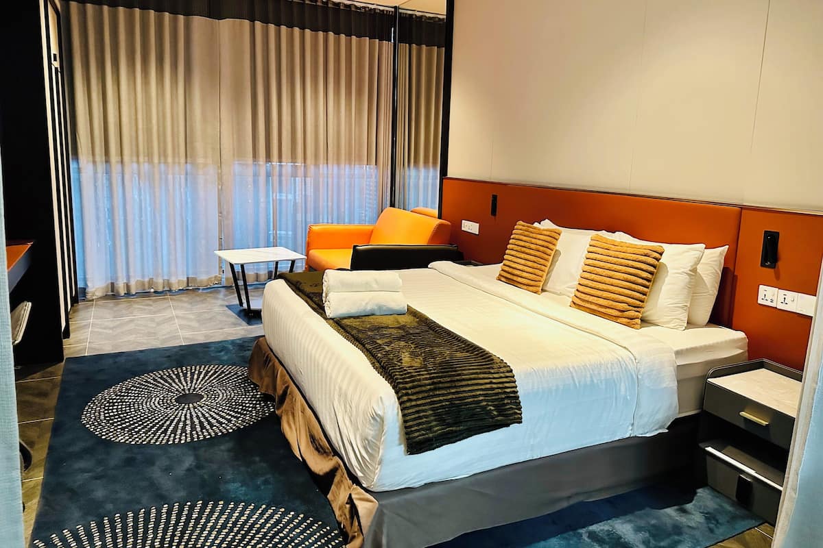 Hotelkamer met een groot bed, twee nachtkastjes, een klein tafeltje, een oranje fauteuil en uitzicht op de stad door dunne gordijnen.
