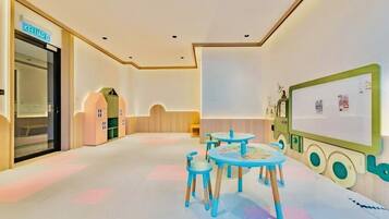 A children's playroom with a table and chairs, a large green screen, and a door labeled "KELUAR".