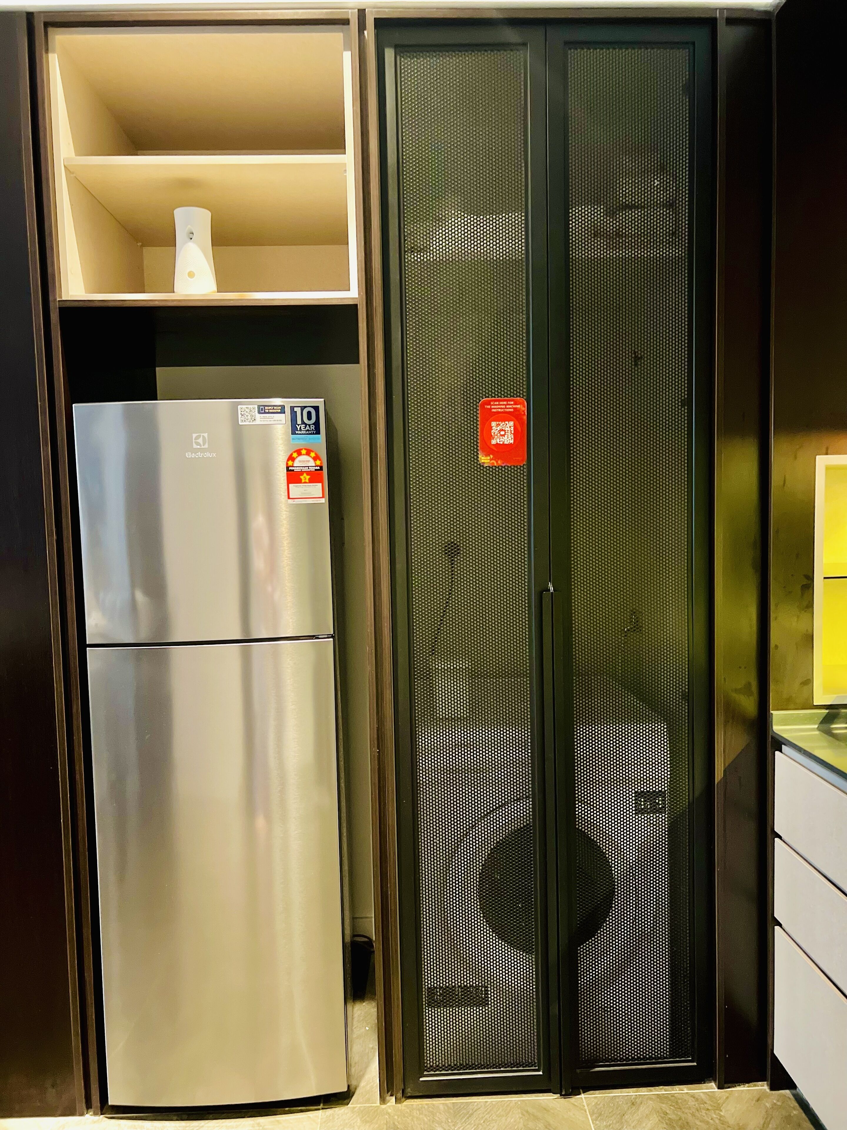 A compact kitchen with a stainless steel refrigerator, a microwave, and a small shelf above the fridge.