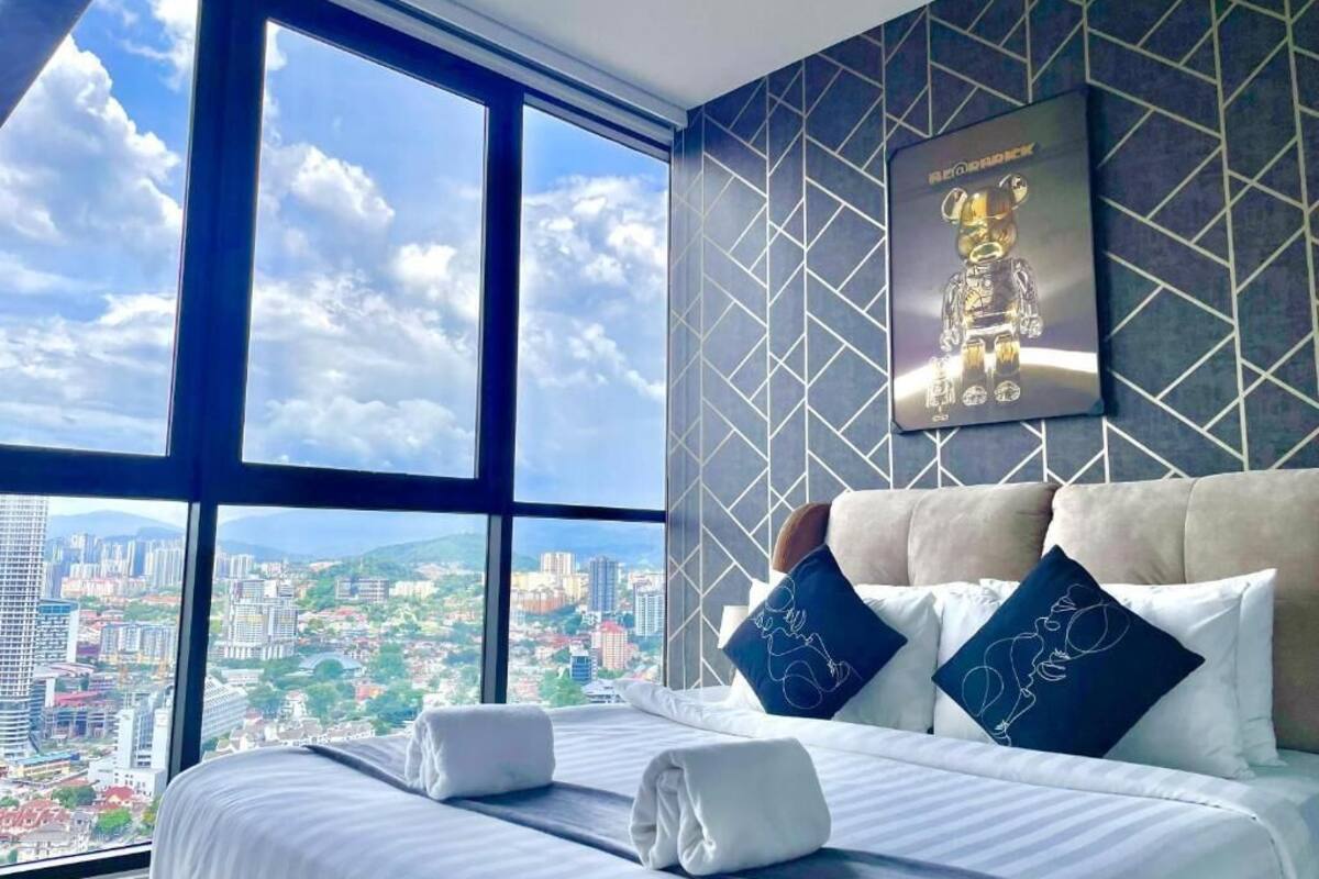 Een hotelkamer met een groot raam met uitzicht op de stad, een netjes opgemaakt bed met kussens en een lijst met een foto aan de muur.