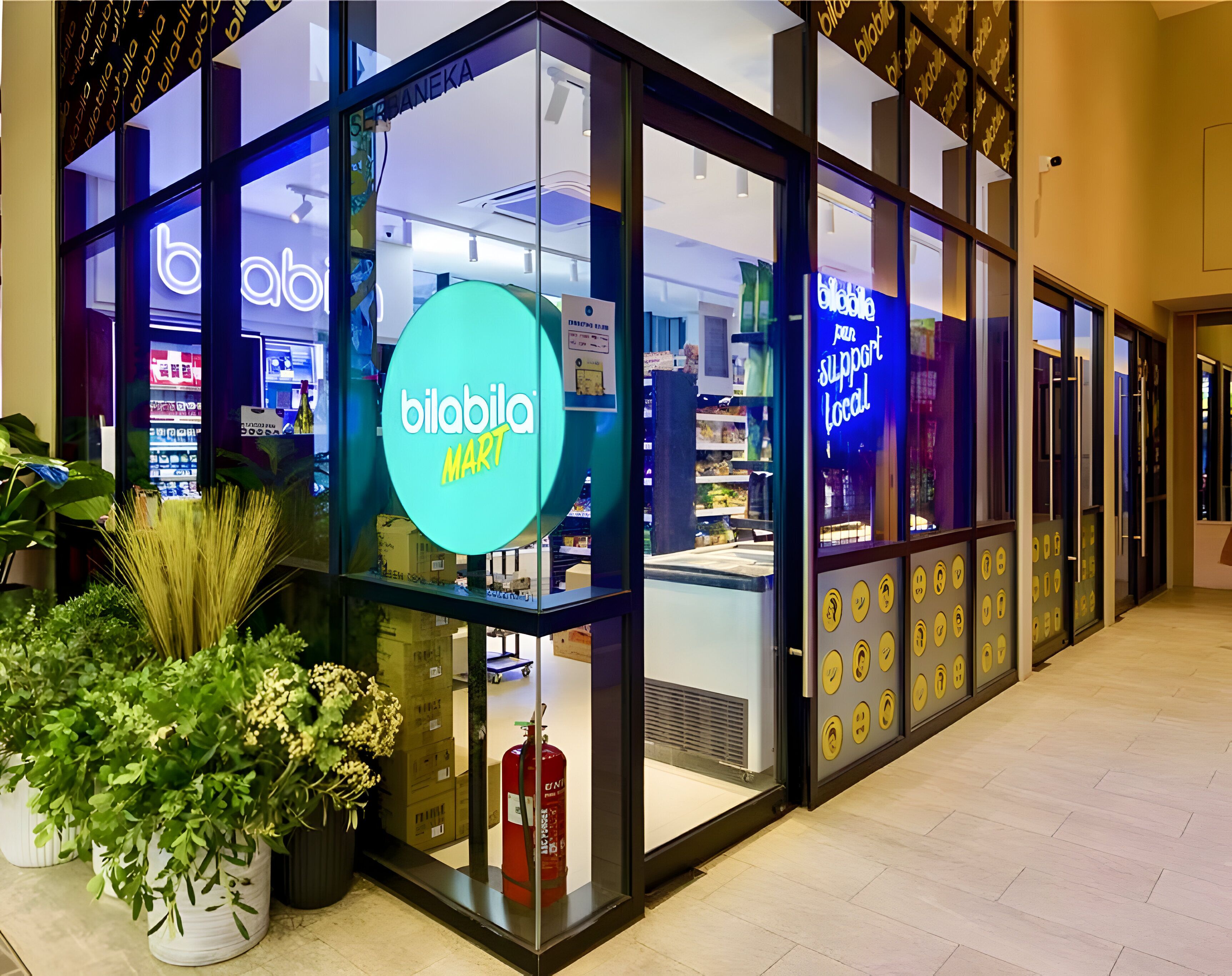 Un magasin appelé Bilabila Mart, avec une grande enseigne lumineuse, des vitrines et des plantes en pot à l’extérieur.