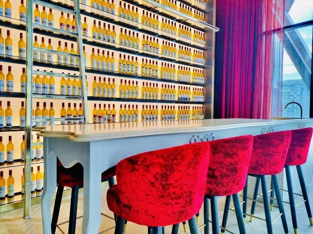 A bar area with a long counter and red bar stools. Behind the counter, there are shelves stocked with bottles.