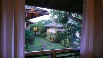 Une vue depuis une fenêtre donnant sur un balcon, avec un jardin bien entretenu en contrebas, comprenant une pelouse, des arbres et une maison en arrière-plan.
