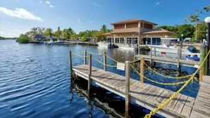 Une jetée avec une passerelle en bois menant à un bâtiment doté d’un toit plat, entouré d’eau et de palmiers.