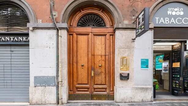 A wooden double door with a metal arch above it, flanked by a closed shutter on the left and a storefront on the right. - BDC - Rome with a view! (Roma)