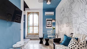 A modern living room with a blue accent wall, a large window, a glass table, a sofa with polka dot pillows, and a wall art piece. - BDC - Rome with a view! (Roma)