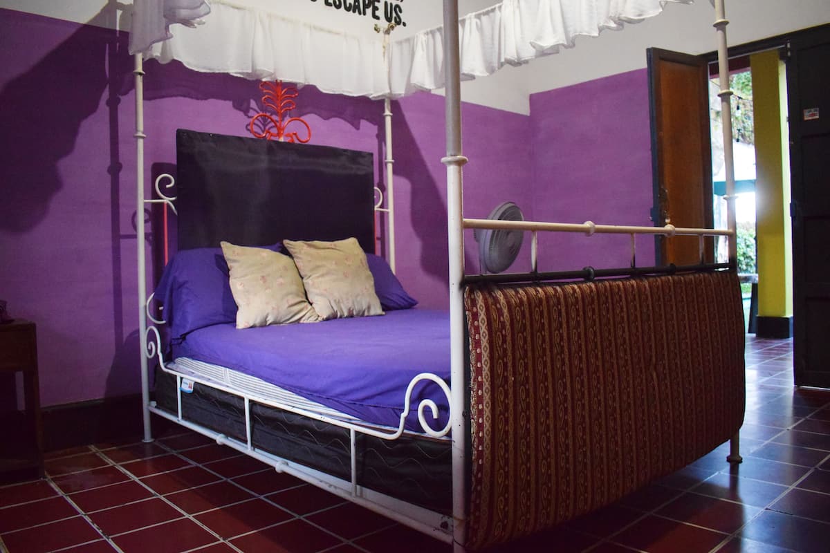 A four-poster bed with a purple bedspread and white pillows, a white canopy, and a red patterned throw blanket.