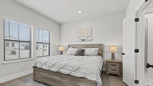 A bedroom with a large bed, two bedside tables with lamps, a door, and a window with blinds. - Brand New Build | Hot Tub | Near Main (Fredericksburg)