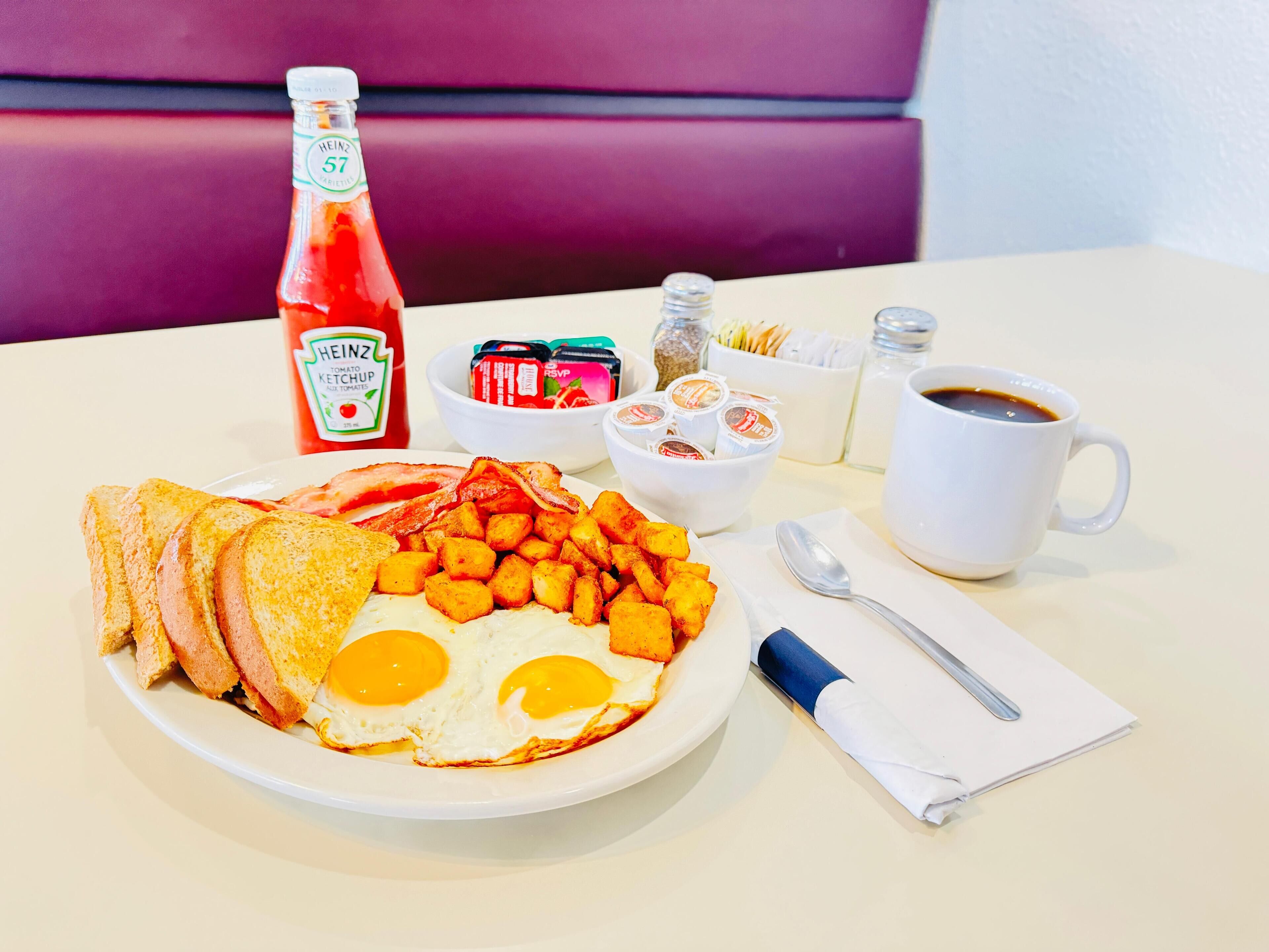 A breakfast plate with eggs, bacon, toast, and potatoes, a bottle of ketchup, condiments, and a cup of coffee on a table.
