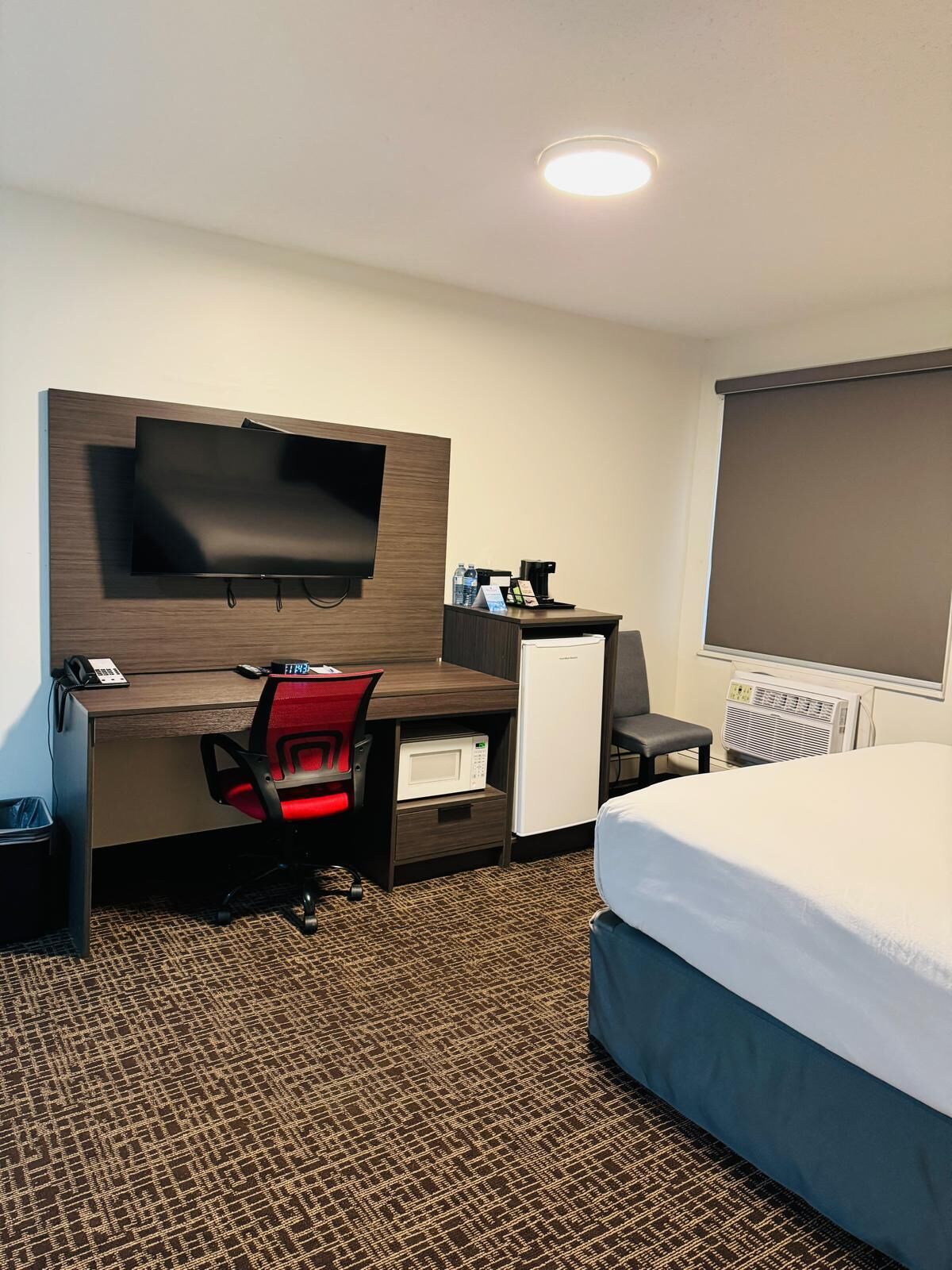 A hotel room with a bed, a desk with a flat-screen TV, a microwave, a coffee maker, and a small refrigerator.
