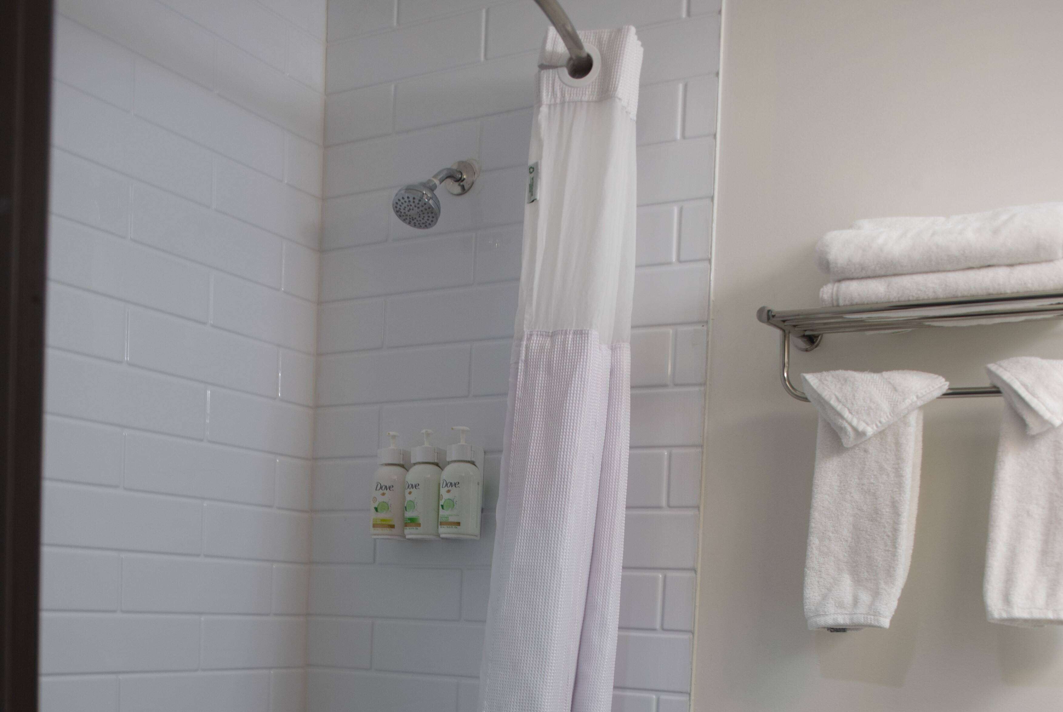 Ducha con cortina blanca, toallero de metal con toallas dobladas y tres botellas de champú Dove en una pared de azulejos blancos.