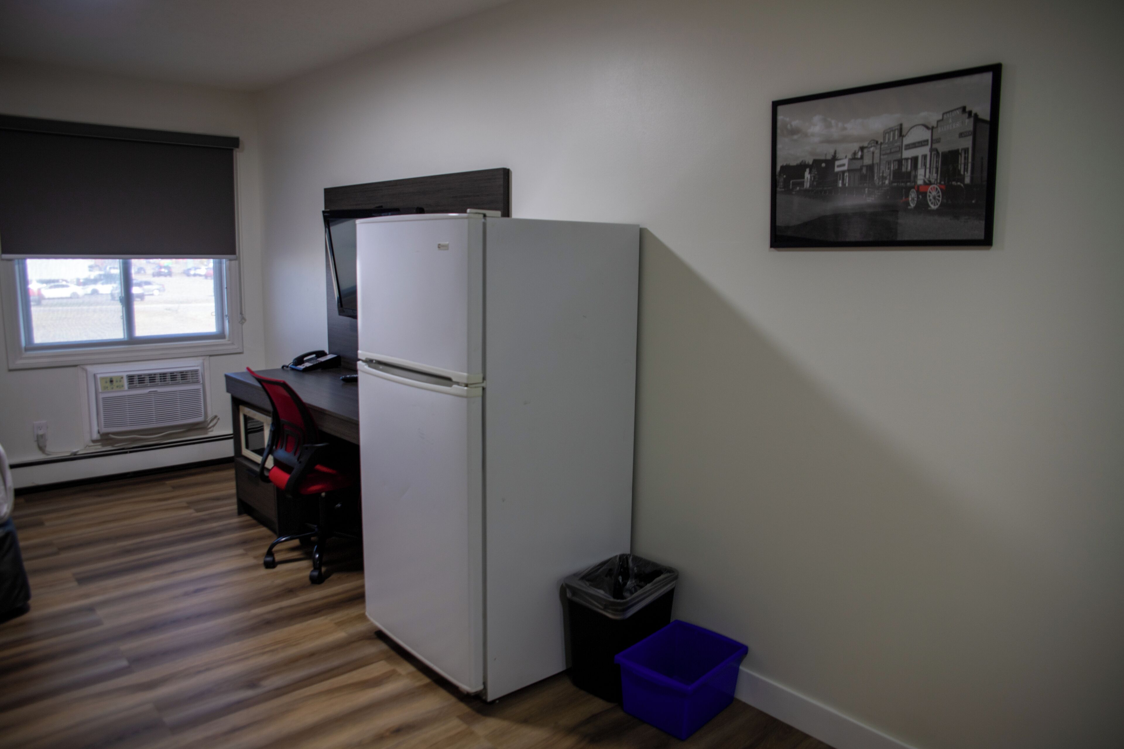 A compact kitchen with a refrigerator, a window with blinds, a small trash bin, and a framed picture on the wall.