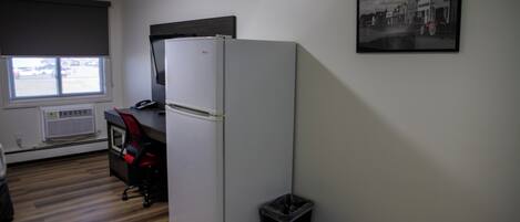 A compact kitchen with a refrigerator, a window with blinds, a small trash bin, and a framed picture on the wall.