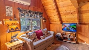 A cozy cabin interior with a wooden ceiling, a sofa with decorative pillows, a television on a wooden stand, and a window overlooking a forest. - Hot Tub in Beautiful Gazebo, Large Covered Outdoor Dining Area, Playground! (Duck Creek Village)