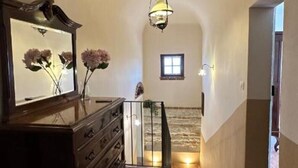 Un hall spacieux doté d’un sol en pierre, d’une commode en bois et d’un miroir qui reflète des fleurs. On peut apercevoir un luminaire suspendu et une petite fenêtre.