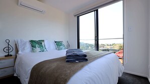 A hotel room with a large bed, white bedding, a view through a large window, and a small bedside table with a decorative item. - Modern 4BR park-view home in St Johns (Auckland)