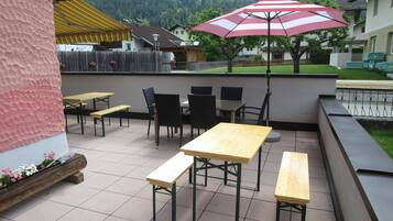Eine Terrassengaststätte mit Holztischen und -stühlen, einem roten und weißen Streifen-Sonnenschirm und Blick auf Häuser im Hintergrund.