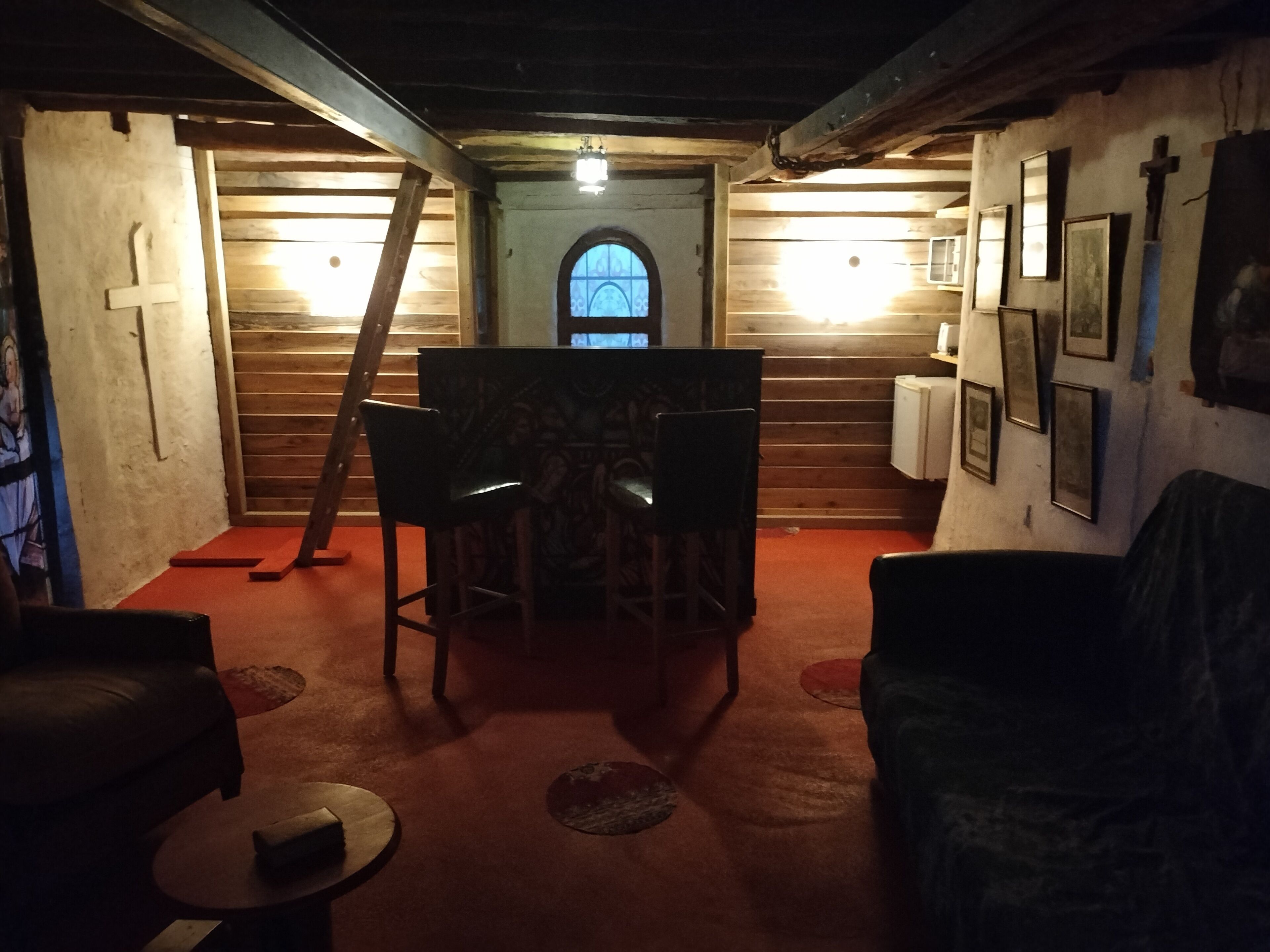 Una habitación tenuemente iluminada con piso de madera, una pequeña mesa, una silla y un sofá. Se ve una escalera apoyada contra la pared y una ventana con una cruz.