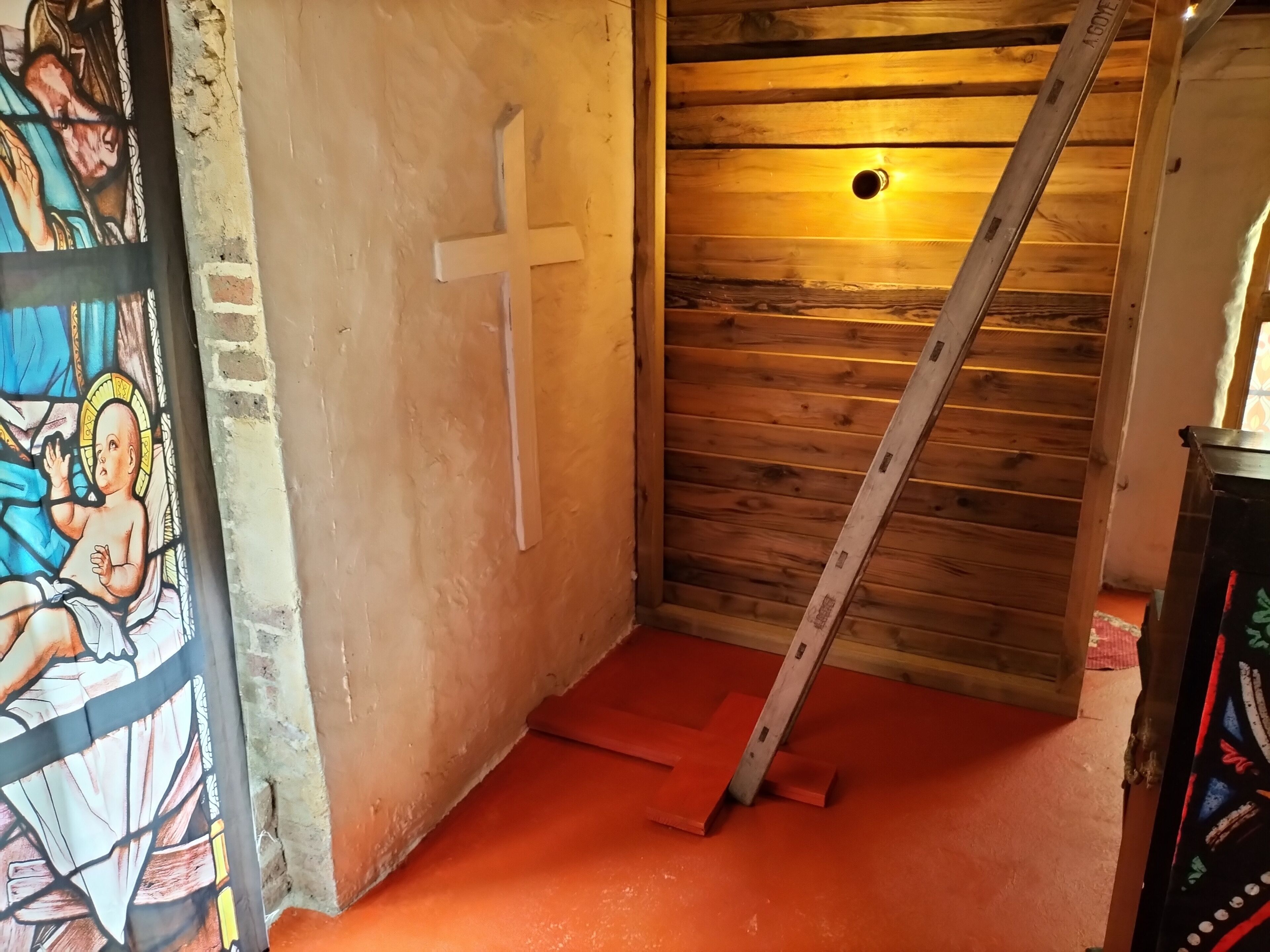 A room with a wooden ceiling, a white cross on the wall, and a ladder leaning against the wall.