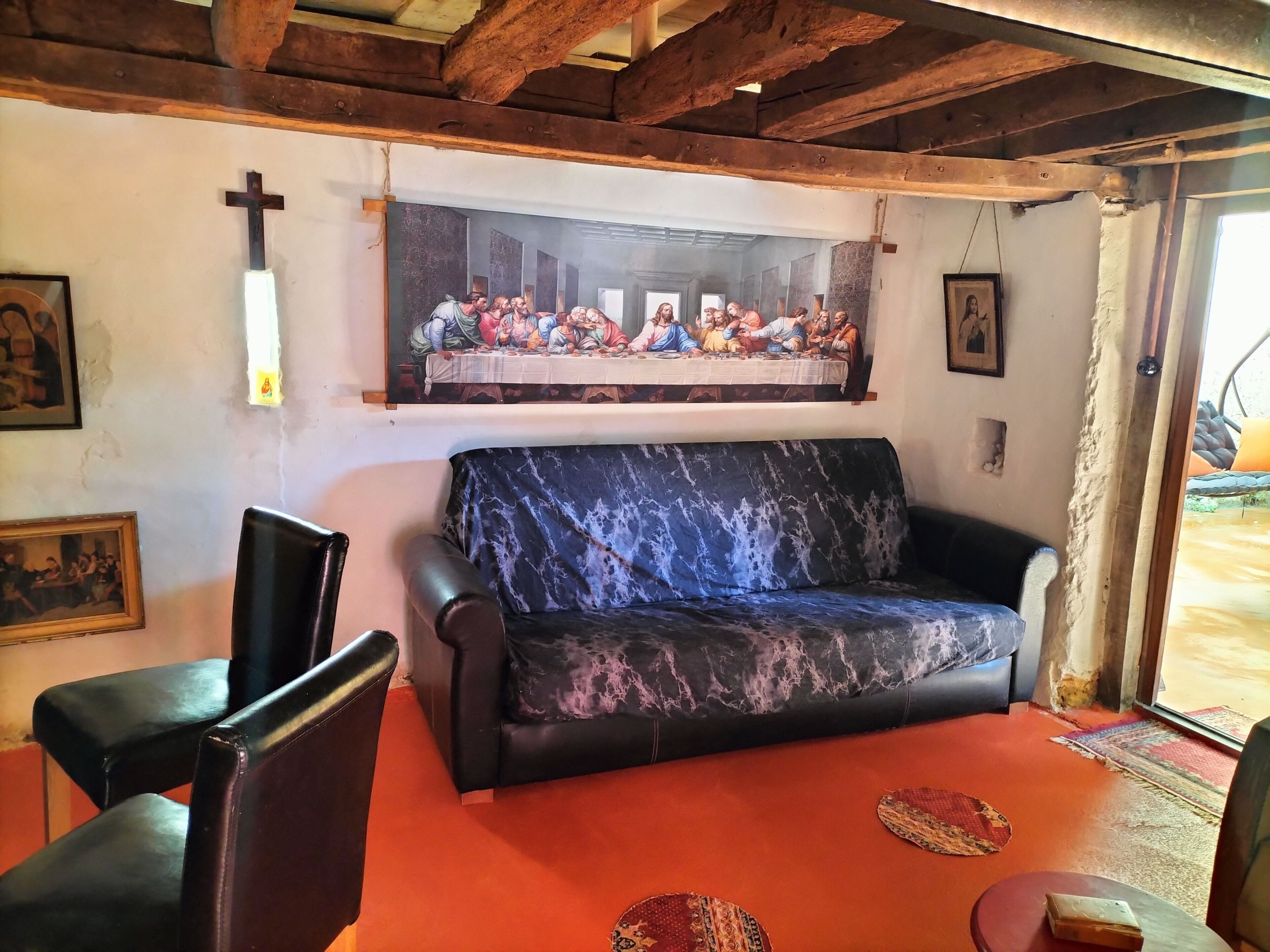Une pièce avec un plafond en bois, un canapé noir, une croix au mur et un tableau représentant « La Cène ».