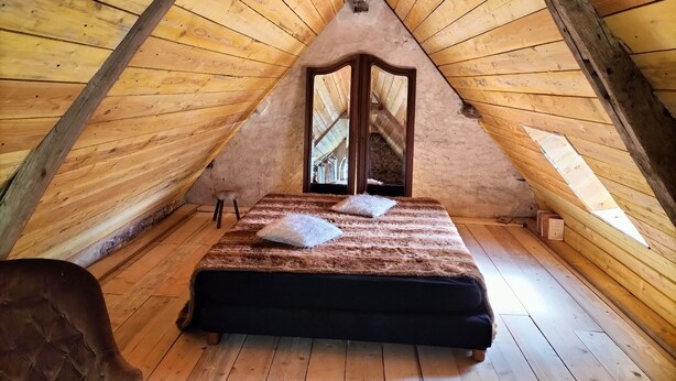 Un dormitorio acogedor en el ático, con techo inclinado de madera, una cama con manta mullida y un taburete pequeño.