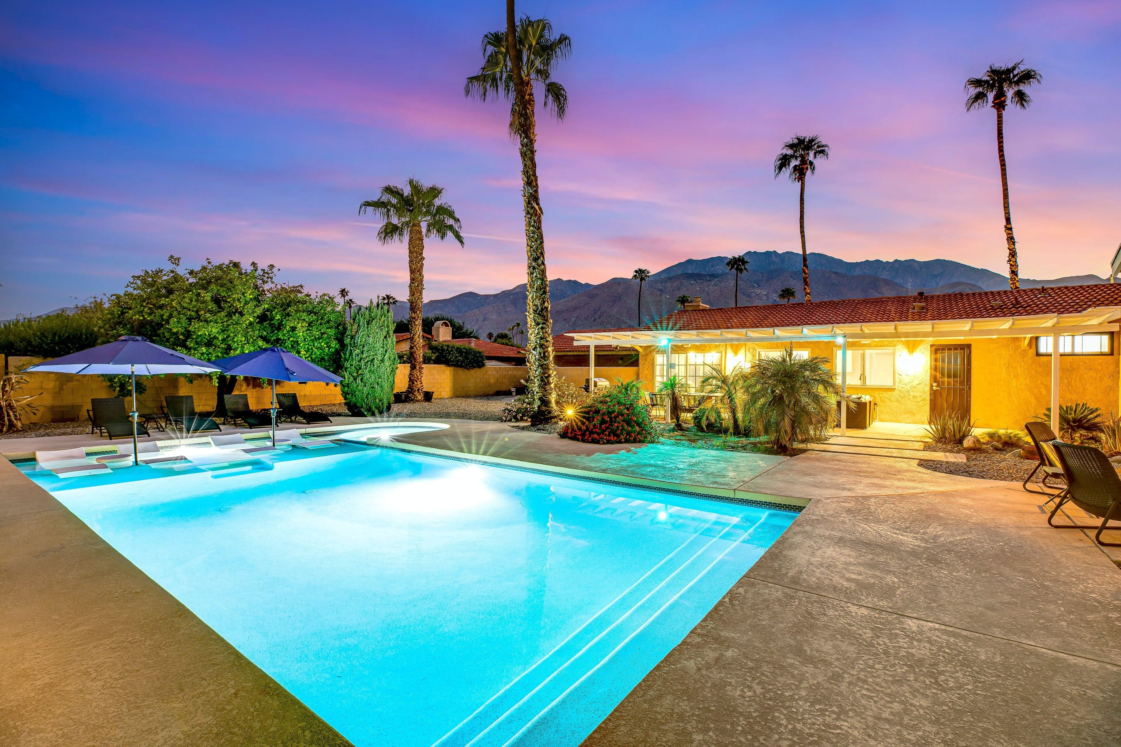 A swimming pool with lounge chairs and umbrellas, a building with a red-tiled roof, and palm trees.