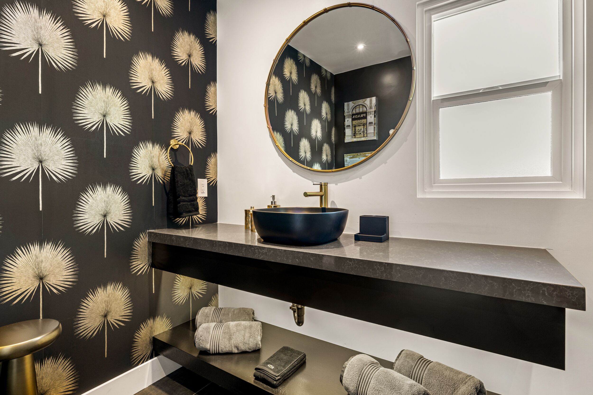 A modern bathroom with a black and white dandelion wallpaper, a round mirror, a dark blue vessel sink, and a black countertop.