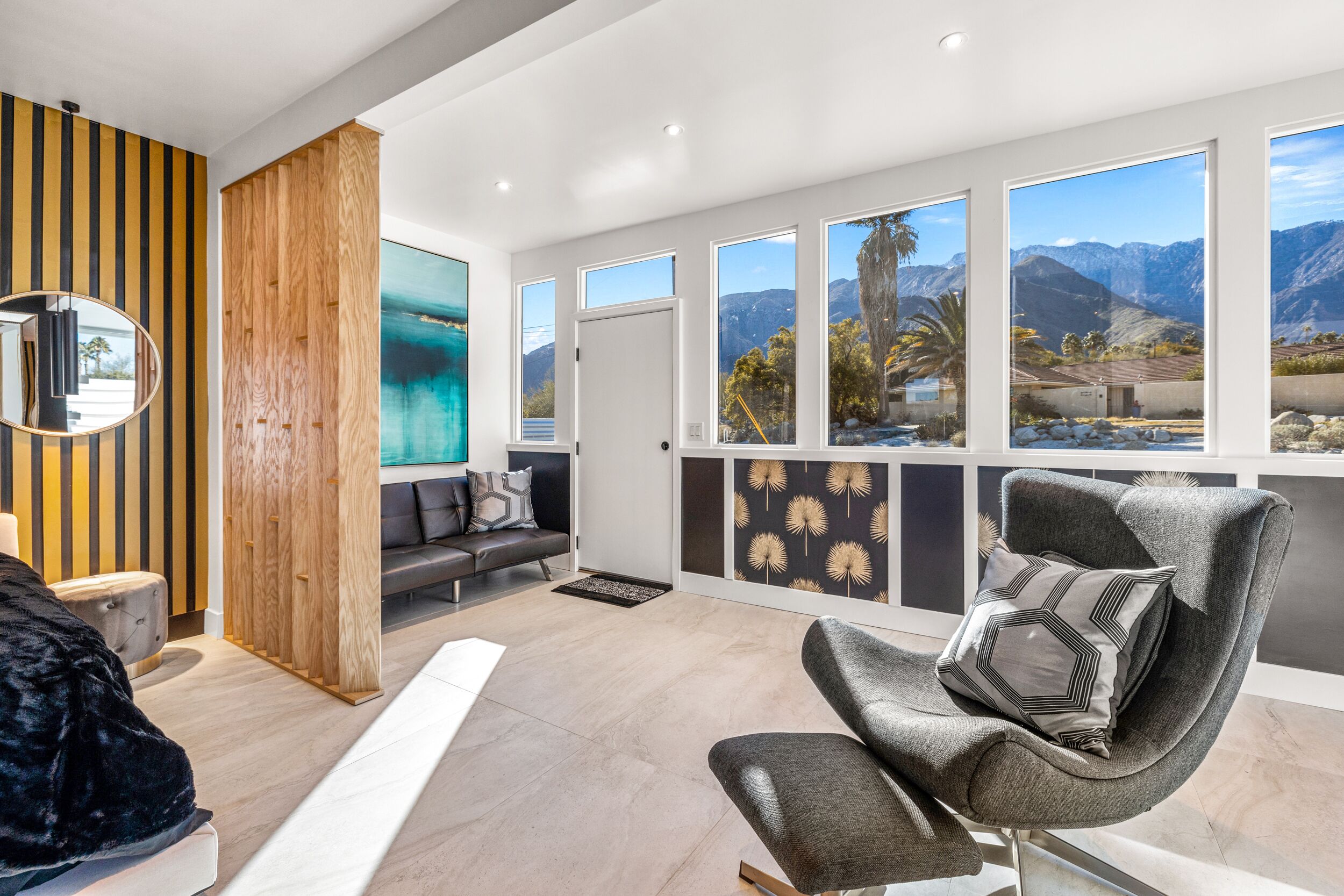 A modern living room with a large window, a grey armchair, a wooden accent wall, and a black sofa.