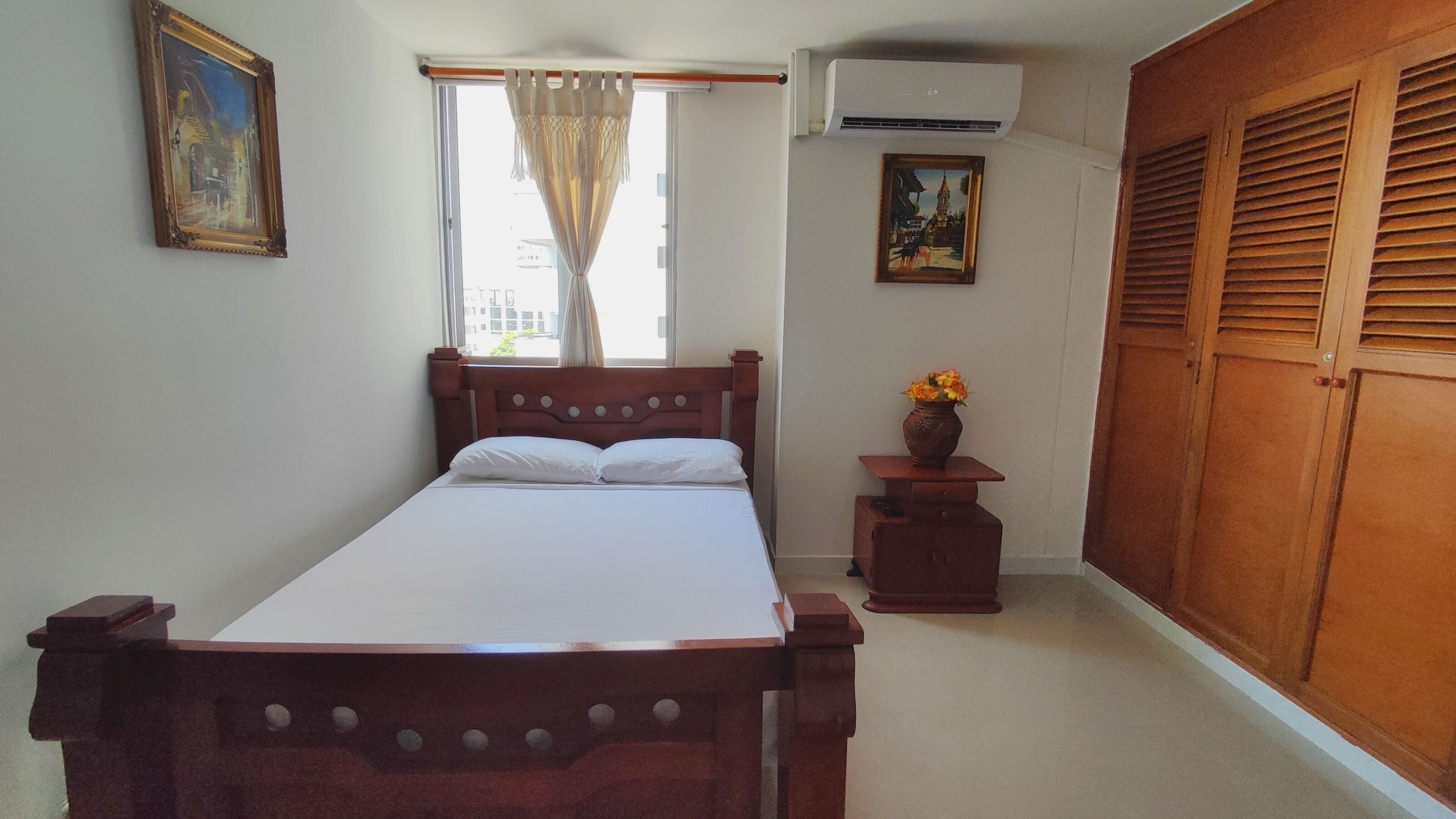 Un dormitorio con cama de madera, una mesita de noche, un florero con flores y un armario.