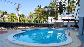 A swimming pool with a tiled deck, surrounded by palm trees and a building in the background. - Beautiful Apartment in Bocagrande  (Cartagena)