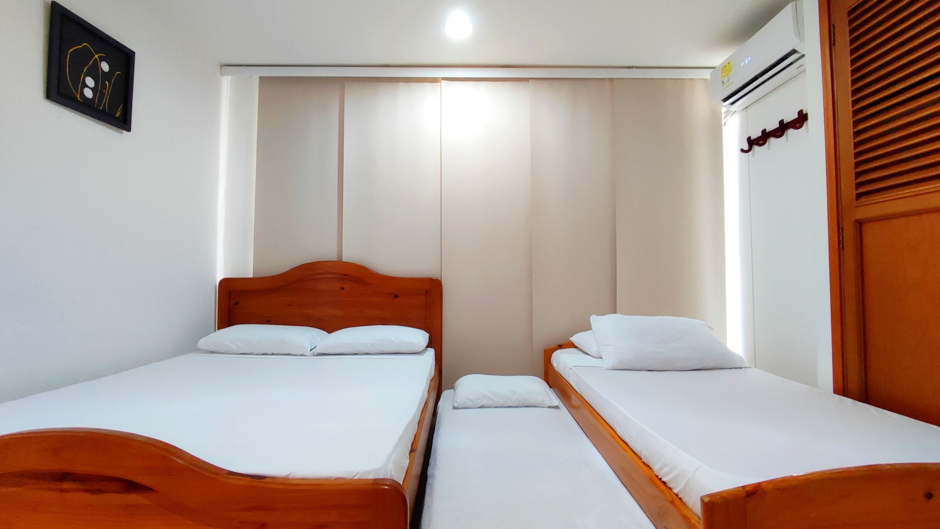 Une chambre avec deux lits en bois, du linge de lit blanc, un tableau encadré au mur et une fenêtre avec des stores.