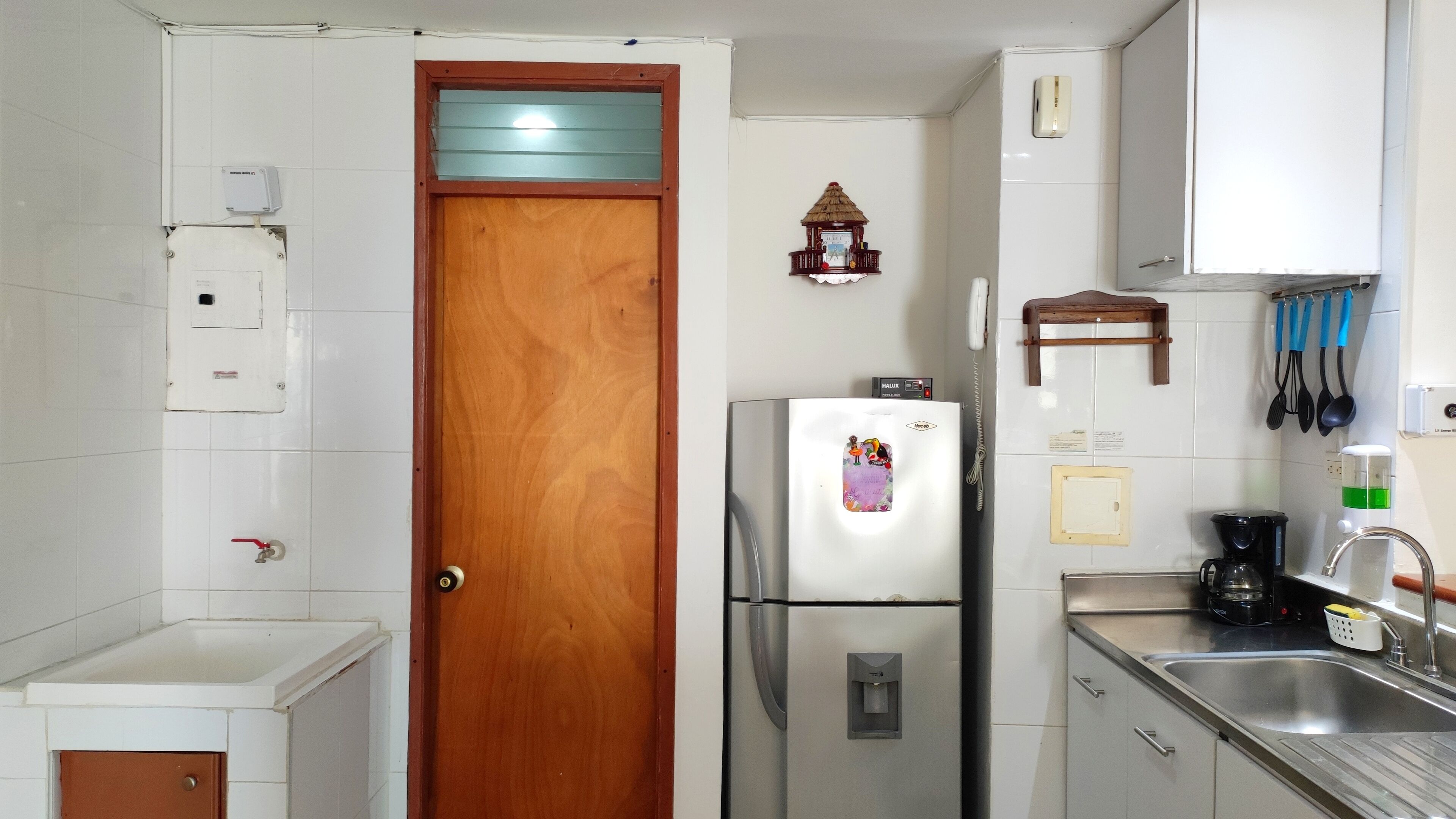 A compact kitchen with a refrigerator, a wooden door, a wall-mounted clock, and basic amenities.