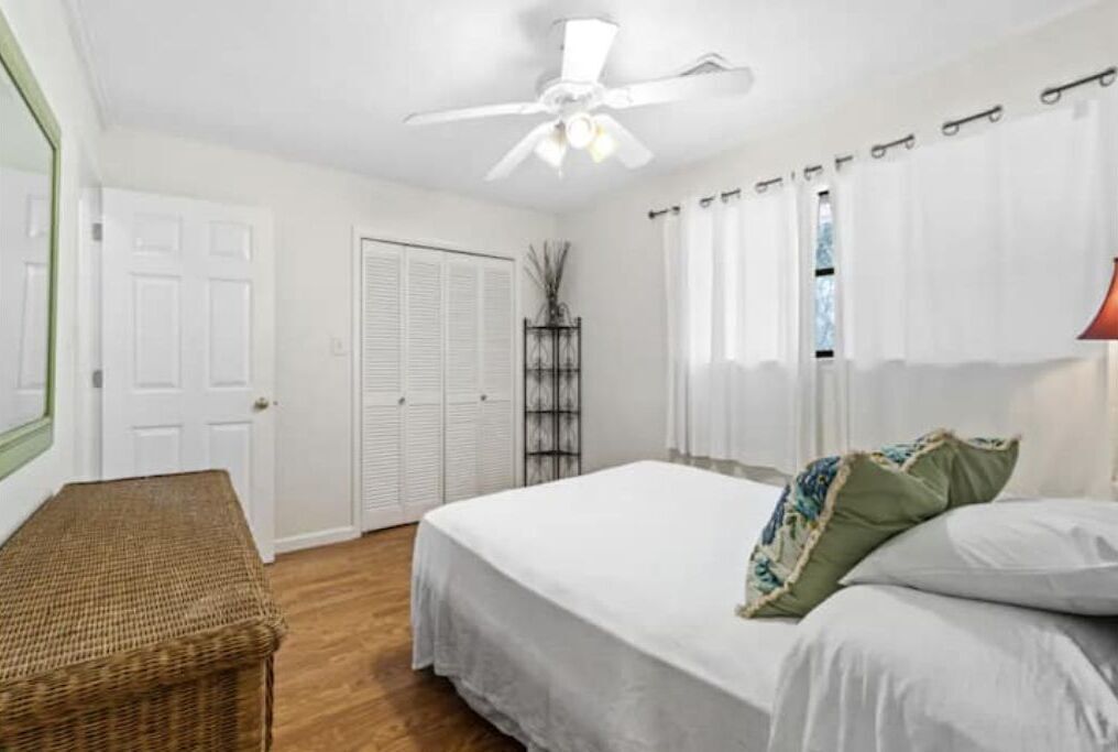 A bedroom with a bed, a ceiling fan, a wicker bench, and a window with curtains.