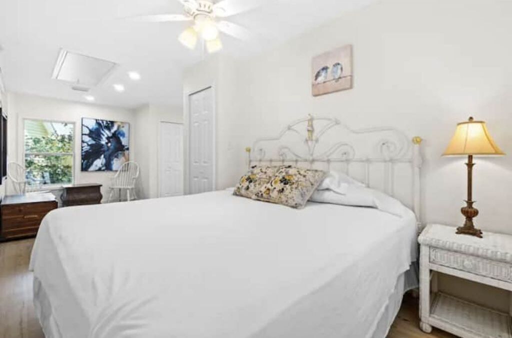 Une chambre à coucher comprenant un lit, une table de chevet, une lampe, un ventilateur de plafond et une fenêtre donnant sur des arbres.