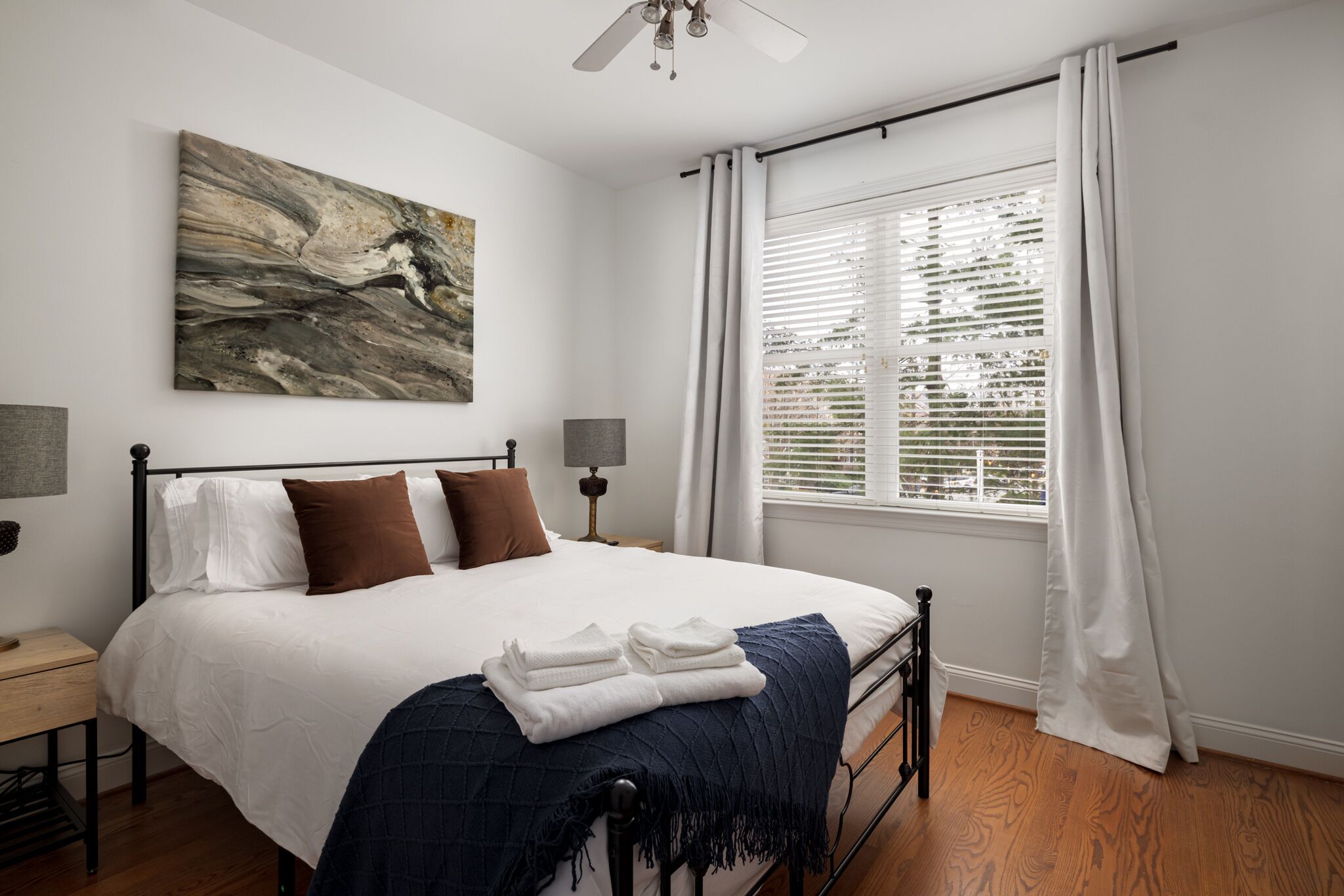 A bedroom with a bed, a ceiling fan, a window with blinds, and a painting on the wall.