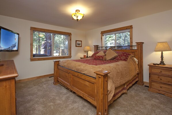 A bedroom with a wooden bed, two windows, a television, a nightstand, and a lamp.