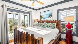 Une chambre avec un cadre de lit en bois, un ventilateur de plafond, une grande fenêtre avec des stores et un tableau sur le thème de la plage accroché au mur.