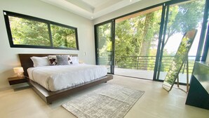 A modern bedroom with a large bed, a nightstand, a bedside table, a floor-to-ceiling sliding glass door, and a view of greenery outside. - Outstanding High End Luxury, Most Convenient Location and World Class Ocean View (Uvita)