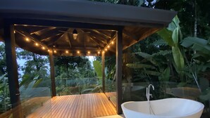 A glass-enclosed outdoor area with a wooden deck, a white freestanding bathtub, and string lights. - Outstanding High End Luxury, Most Convenient Location and World Class Ocean View (Uvita)