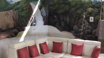 Un sofá modular blanco con cojines rojos, una mesa de centro negra y un mural de una cascada y un bosque.