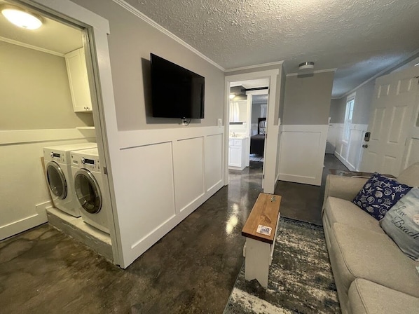 A living room with a beige sofa, a coffee table, and a flat-screen TV mounted on the wall. There is a washing machine in the adjacent laundry room. - Cottage on the Marsh - Walk to Beach in Garden City, SC (Garden City Beach)