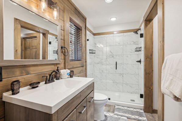 Un baño moderno con tocador de madera, un espejo grande, un lavamanos blanco, inodoro y ducha a ras de suelo con azulejos de mármol.