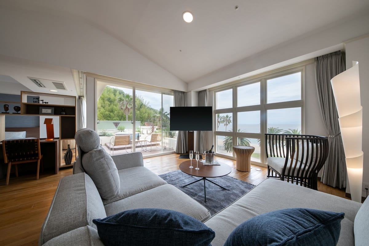 A modern living room with a large sofa, a TV, and a balcony with a view of the sea.