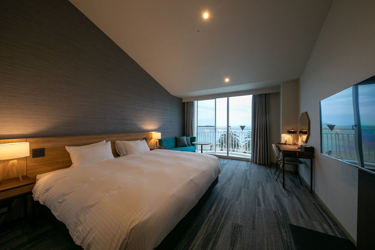 A hotel room with a large bed, a desk with a chair, a small table, and a balcony with a view.