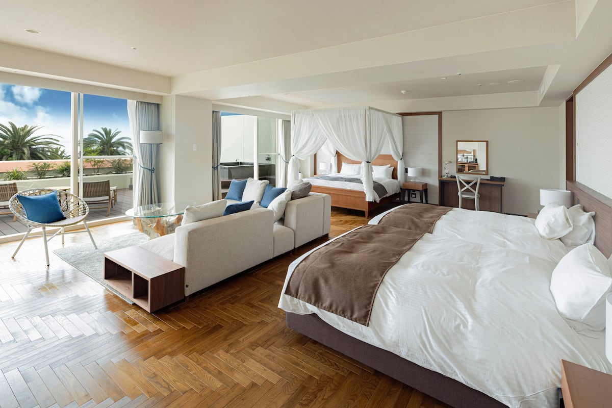 A spacious bedroom with a large bed, a sofa, a desk, and a balcony with a view of palm trees.