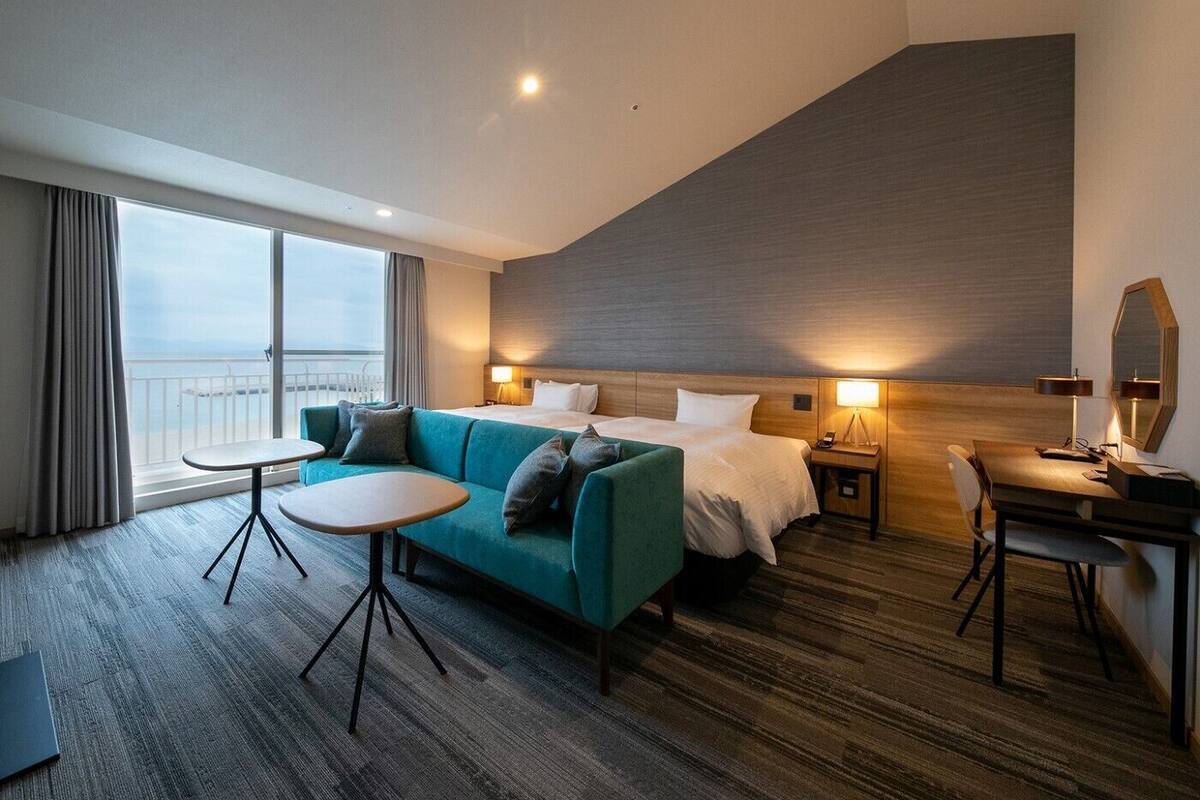 A modern hotel room with a large bed, a sofa, a small table, a desk, and a view of the sea.