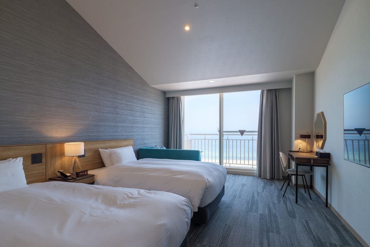 A hotel room with two beds, a desk, a chair, a nightstand, a lamp, and a large window with a view.