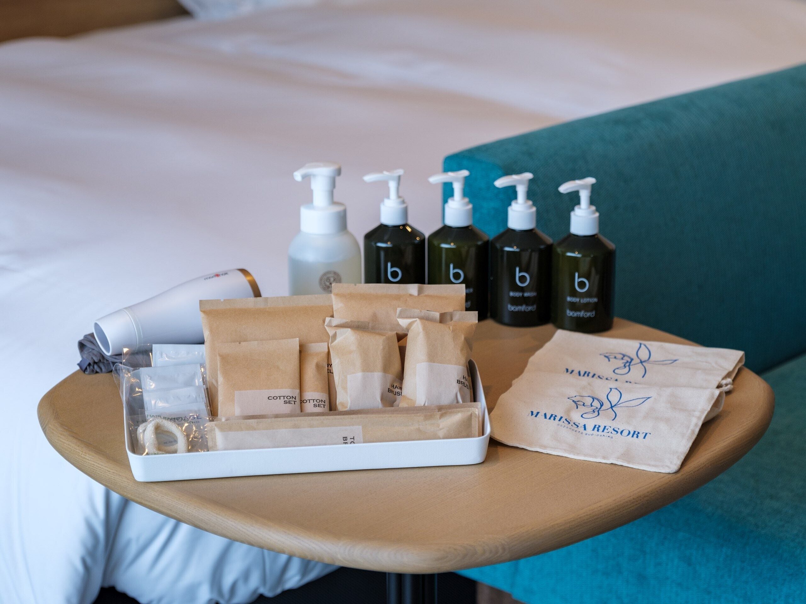 Et pænt anrettet bord med toiletartikler, herunder vatrondeller og flasker med bodylotion, samt en mærkevarepose på et bord på et hotelværelse.