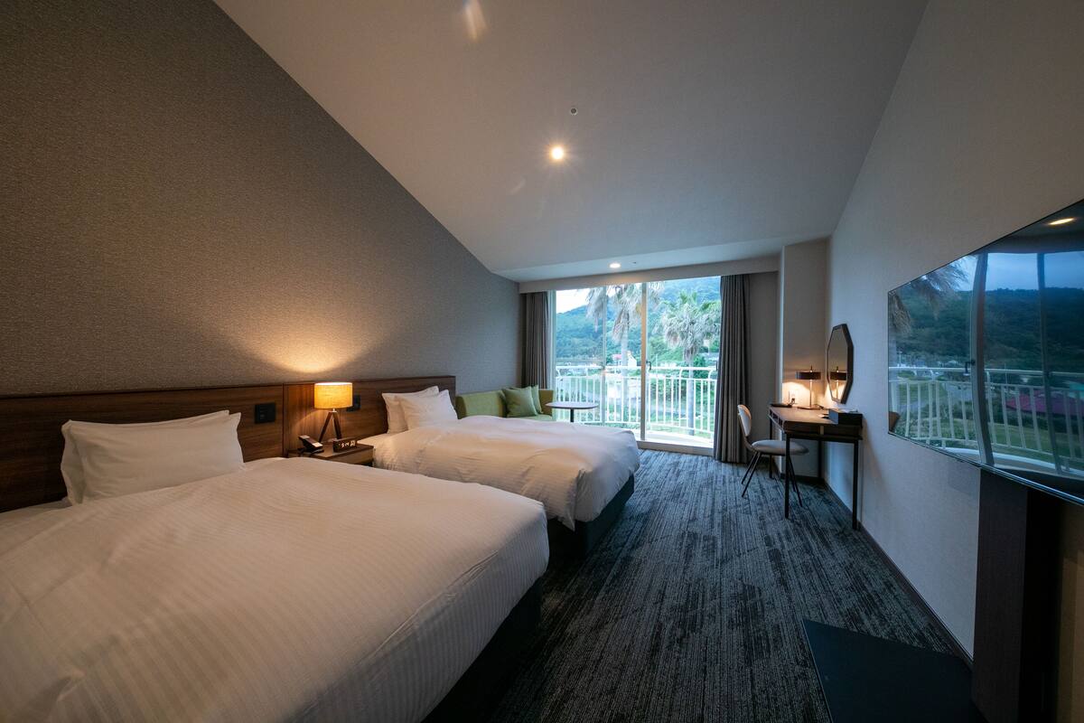 A hotel room with two beds, a desk, a chair, and a large window offering a view of palm trees and a balcony.