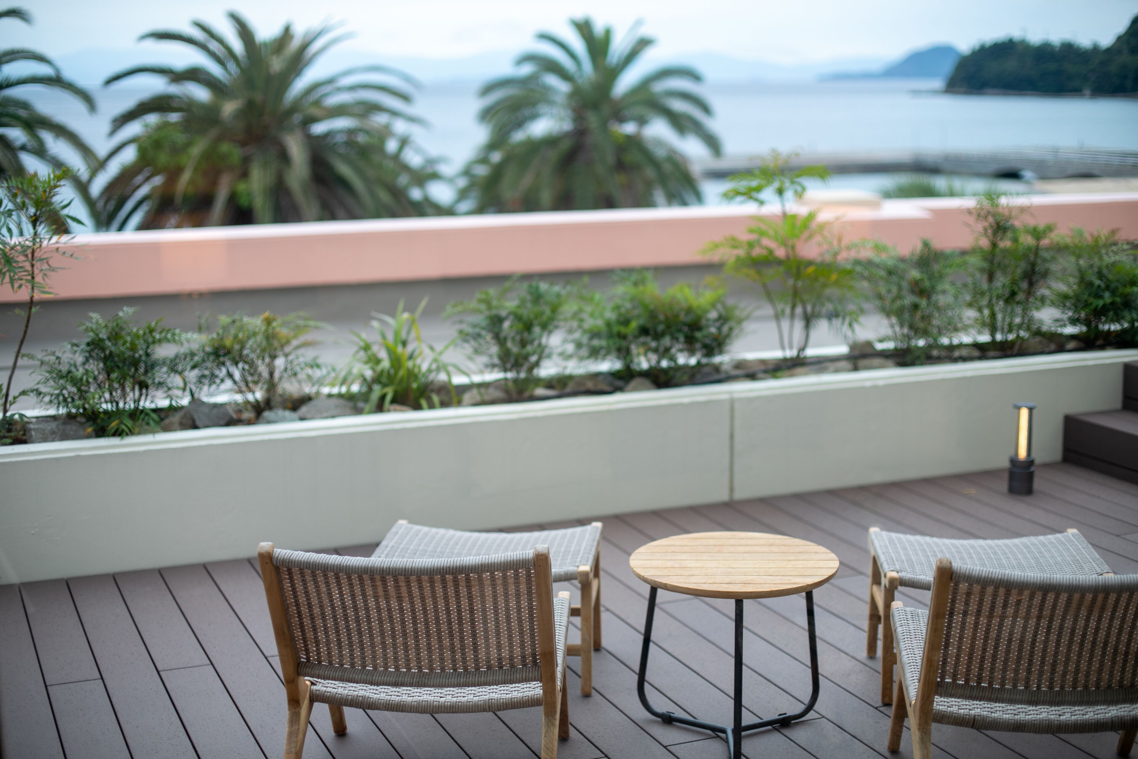 A deck with wicker chairs and a round table, overlooking a landscaped area with palm trees and a view of the sea.